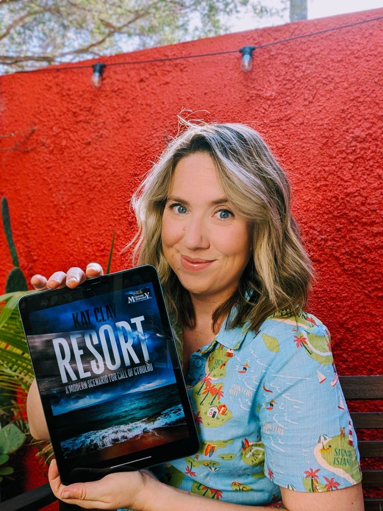 A blonde white woman in a tropical shirt in front of a red wall holding an ipad with the cover of her game, Resort. The cover has an ominous tropical island on it. 