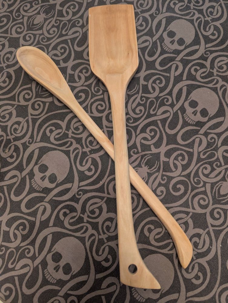 Two long handled wooden kitchen instruments, one is a spoon and the other a wide flat spoonular paddle, on a skull patterned tablecloth.