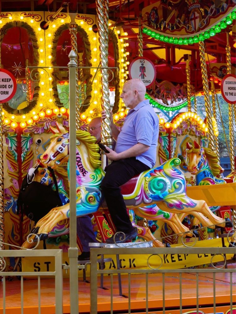 Hochkantfoto: Weißhaariger Mann auf einem quietschbunten Karussellpferd in Seitenansicht. Er trägt ein fliederfarbenes Polo-Shirt und eine schwarze Hose, sein Blick ist ernst nach vorne gerichtet. Das Karussell ist mit zahlreichen bunten Lichterketten geschmückt.