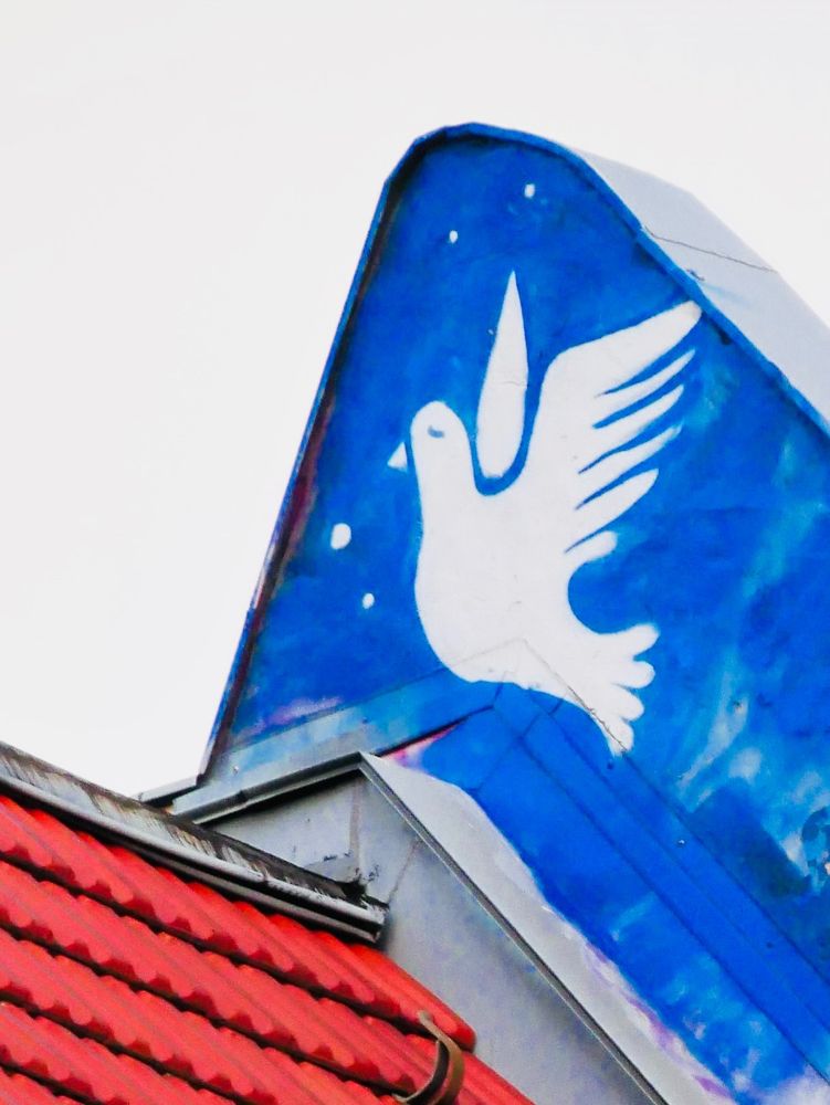 Hochkant-Foto: Ein Dachgiebel, auf dem auf blauem Grund eine weiße Friedenstaube gemalt ist.