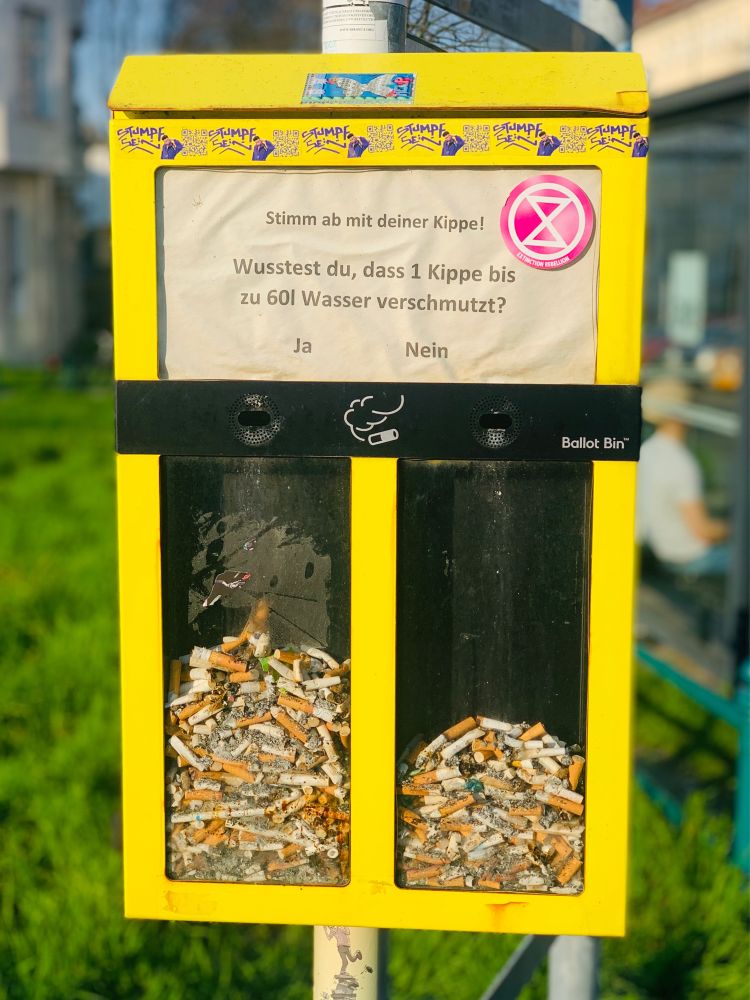 Foto im Hochformat: Gelber Abstimmungskasten, der an einer Stange eines Straßen Schildes befestigt ist. Der Text auf dem Kasten: “Stimm ab mit deiner Kippe! Wusstest du, dass 1 Kippe bis zu 60l Wasser verschmutzt?” Darunter zwei Einwurflöcher mit Ja und Nein. Bei ja ist die Hälfte mit Kippenresten gefüllt, bei Nein ca. ein Drittel. Im Hintergrund ist eine saftiggrüne Wiese zu erkennen.