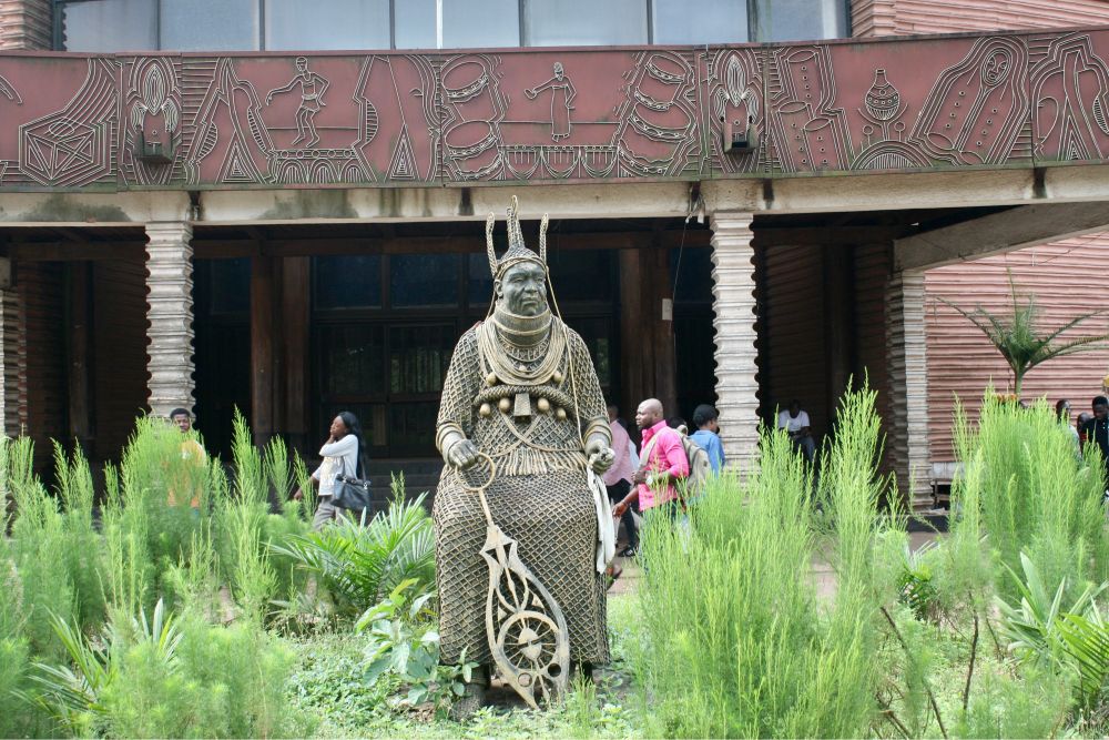 Foto im Querformat: Skulptur eines sitzenden Oba (König) in Korallen gekleidet inmitten grüner Gewächse.