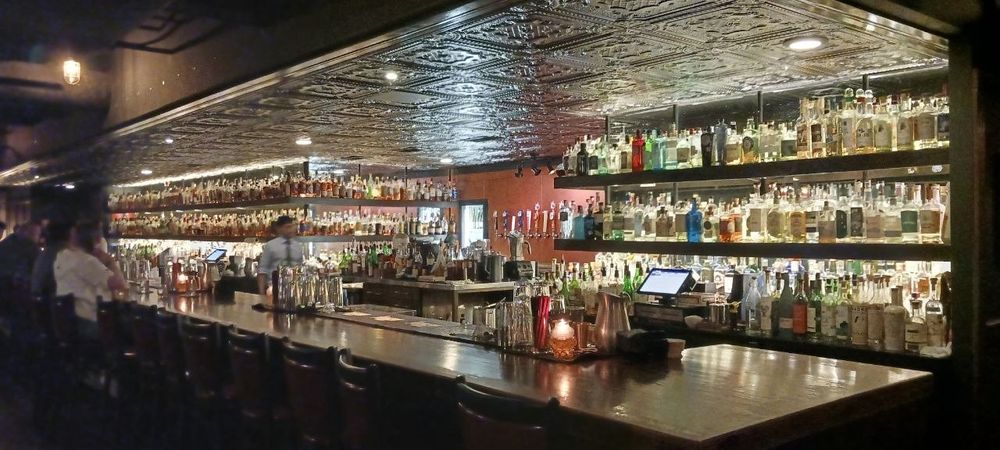 A photo of Bar 1919's bar-top, from the corner. A dimly lit interior with brightly back-lit bar shelves, providing enough light for patrons to see while also being a comfortable level of darkness for privacy, befitting the later hours most patrons arrive at.