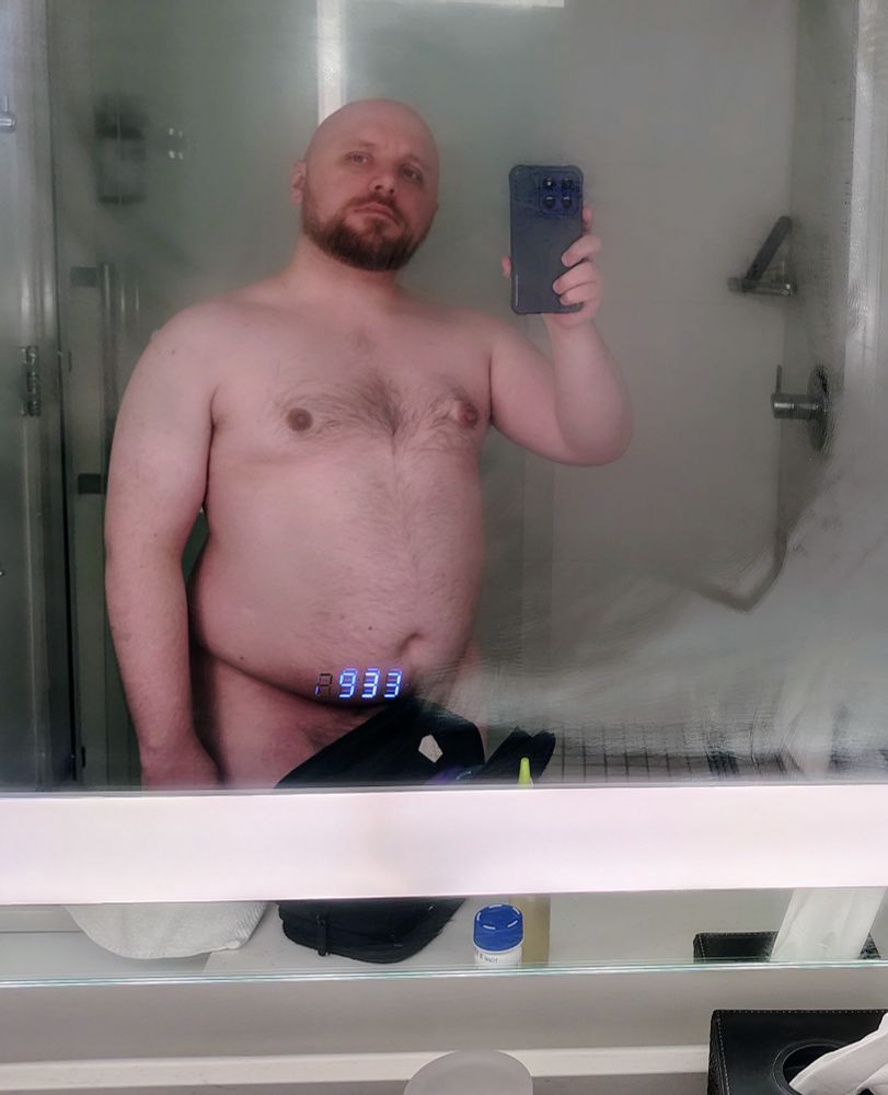Depicted is a bald and bearded man from mid-thigh up. He is taking a selfie in front of a foggy bathroom mirror. Naughty bits are hidden by his travel kit, but this is the only thing covering his body.