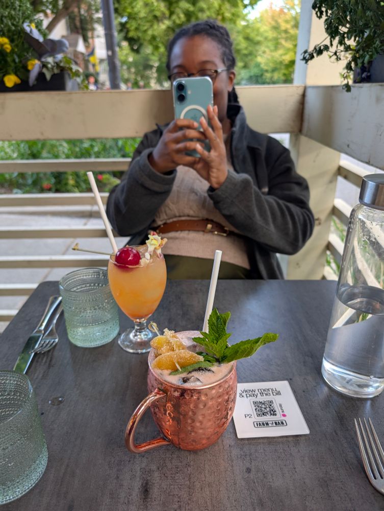 Two fresh cocktails sitting on a wooden table. A Black woman on the other side of the table is taking a photo of the cocktails and the person taking this photo.