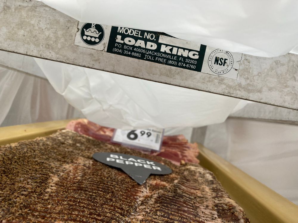 A photo of a bread/bun rack with a label that shows it's of the brand "Load King." It is covered in a plastic sheet, and the first rack visible carries a bunch of black pepper bacon and apple wood smoked bacon.