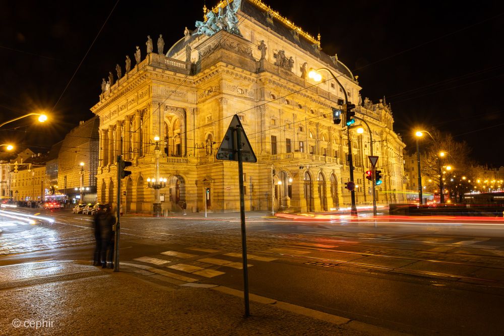 Langzeitbelichtung bei Nacht. Im Zentrum ist ein mit gelblichem Licht beleuchtetes historisches Gebäude zu sehen, das sich an einer Kreuzung befindet. Vorne rechts sind verwischt zwei Fußgänger zu sehen, die an einer Anpel warten. Auf der Kreuzung sind die Lichtspuren von Autos zu erkennen.