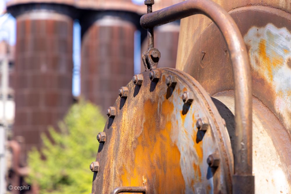 Das Bild zeigt ein verrostetes, industrielles Objekt im Vordergrund, wahrscheinlich Teil einer Maschine. Es hat eine runde Form und ist mit vielen Bolzen versehen. Im Hintergrund sind unscharf große Industriebehälter zu sehen, die ebenfalls einen rostigen oder gealterten Eindruck machen. Das Bild zeigt eine verlassene  Industrieanlage darstellen.
