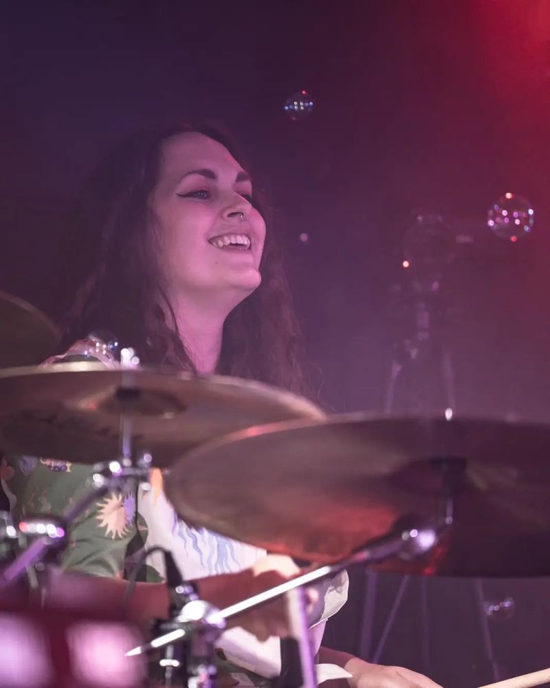 Photo of EBB drummer Anna Fraser, behind her drumkit, playing her little heart out and grinning from ear to ear!