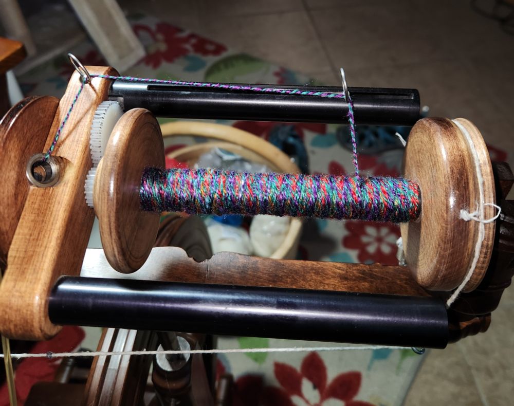 Close up of the Woolee Winder bobbin/flyer on a Kromski Sonata. The yarn is very brightly and multi-colored. Because of the way the Woolee Winder loads the thread, it is impossible to pick out more than glimpses of the red, purple, copper and green. background is a floral rug over a tile floor, and a partially seen basket of fiber