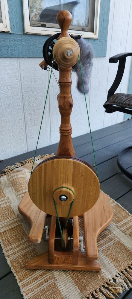 Majacraft Little Gem spinning wheel as seen from the non-treadle side. It is sitting on a tan and beige rug, and there is a tuft of gray metallic fiber visible hanging from the bobbin. The wheel is particularly remarkable for it's very small drive wheel and different style of drive and tension bands.
