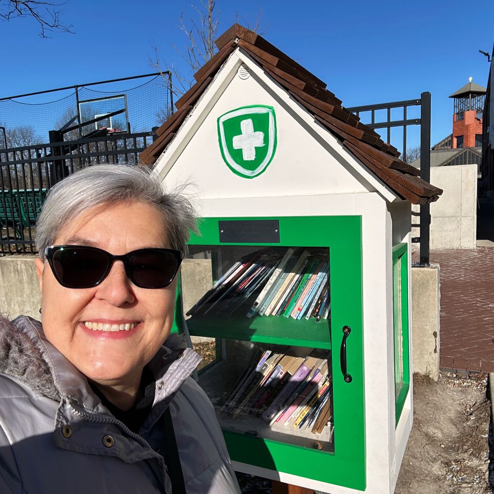 Chris Wolak a white woman with grey hair and sunglasses stands before a white and green Little Free Library