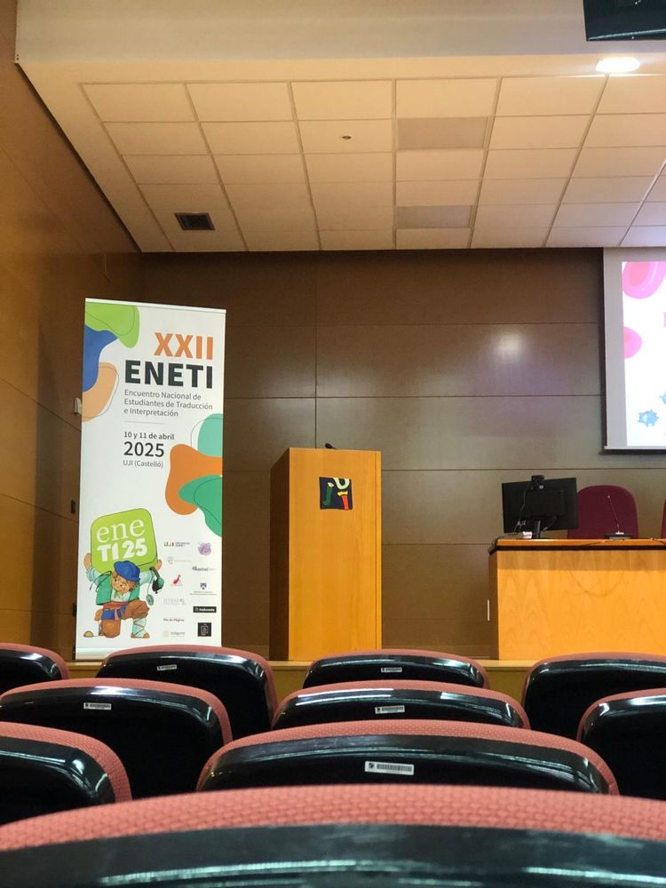 Vista del escenario del salon de actos de la facultad de jurídicas de la Universidad Jaume I de Castellón. A la izquierda, un cartel del XXII ENETI; en el centro, un atril; a la derecha, la mesa de ponencias. 