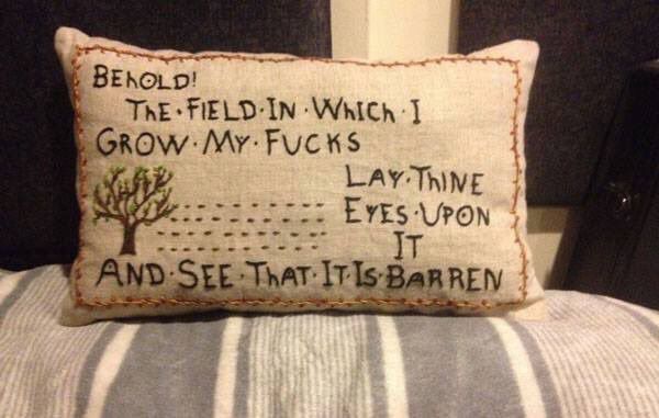 An embroidered pillow reading BEHOLD! The field in which I grow my fucks. Lay thine eyes upon it and see that it is barren