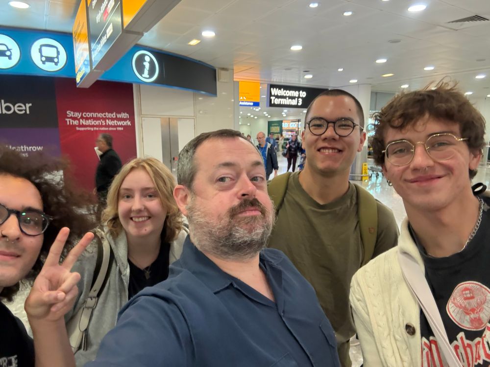 Five people, including me, at Heathrow Airport arrivals. It’s a selfie, I’m taking it, and we’re all looking at the camera and smiling. 