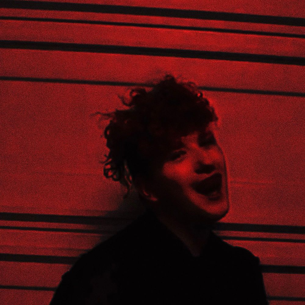 Heavily edited black and red photo of the artist smiling incredibly wide with demon fangs 