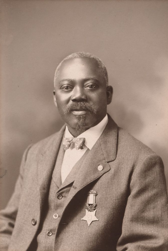 Of the 3,498 service members who have received the Medal of Honor throughout U.S. history, only 88 have been black. The first, pictured here in a black and white photo is Sgt William Carney shown wearing a 3 piece suit and his medal of honor pinned to his left chest pocket area. 
On July 18, 1863, the soldiers of Carney's regiment led the charge on Fort Wagner. During the battle, the unit's color guard was shot. Carney, who was just a few feet away, saw the dying man stumble, and he scrambled to catch the falling flag.

Despite suffering several serious gunshot wounds himself, Carney kept the symbol of the Union held high as he crawled up the hill to the walls of Fort Wagner, urging his fellow troops to follow him. He planted the flag in the sand at the base of the fort and held it upright until his near-lifeless body was rescued.

Even then, though, he didn't give it up. Many witnesses said Carney refused to give the flag to his rescuers, holding onto it tighter until, with assistance, he made it to the Union's temporary barracks. 

Promoted for His Actions
Carney lost a lot of blood and nearly lost his life, but not once did he allow the flag to touch the ground. His heroics inspired other soldiers that day and were crucial to the North securing victory at Fort Wagner. Carney was promoted to the rank of sergeant for his actions.

For his bravery, Carney was awarded the Medal of Honor on May 23, 1900.