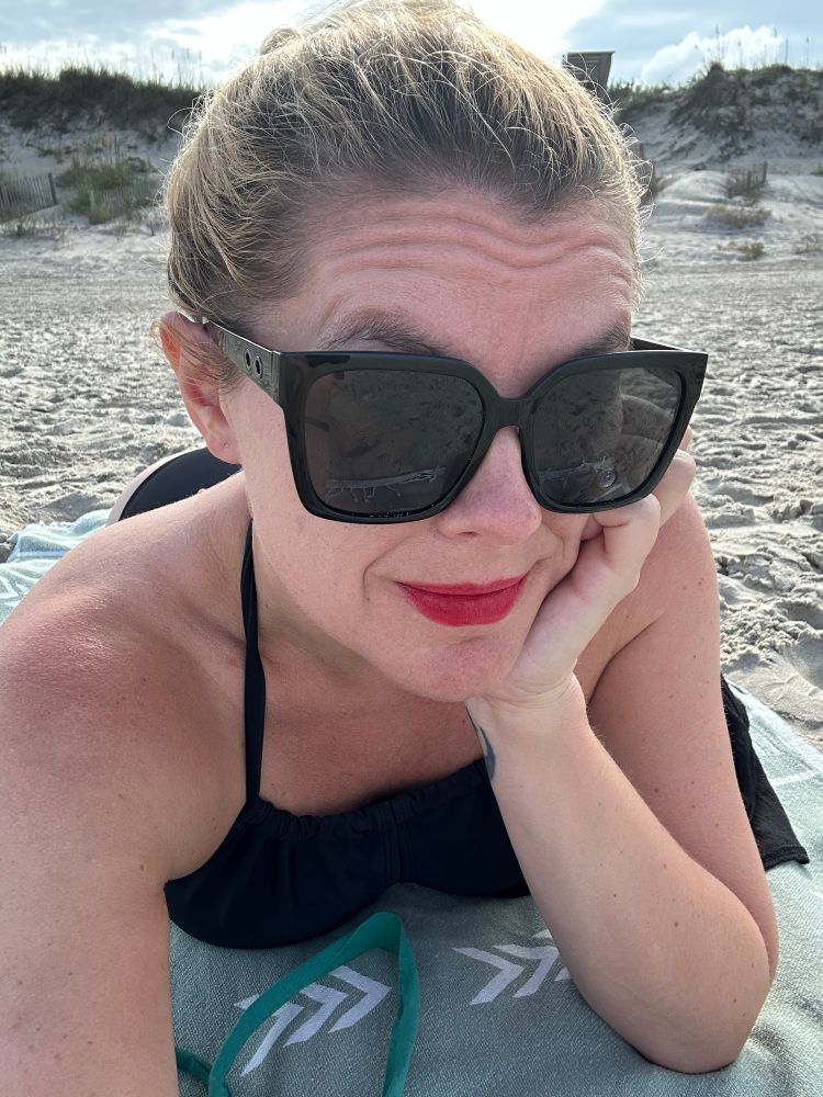 Cass stretched out on her stomach on a pale green beach towel, resting her chin on one hand. Dhe’s wearing a black halter top swimsuit, black sunglasses, and red lipstick, with her hair pulled up in a messy knot. Sand dunes are visible in the background.