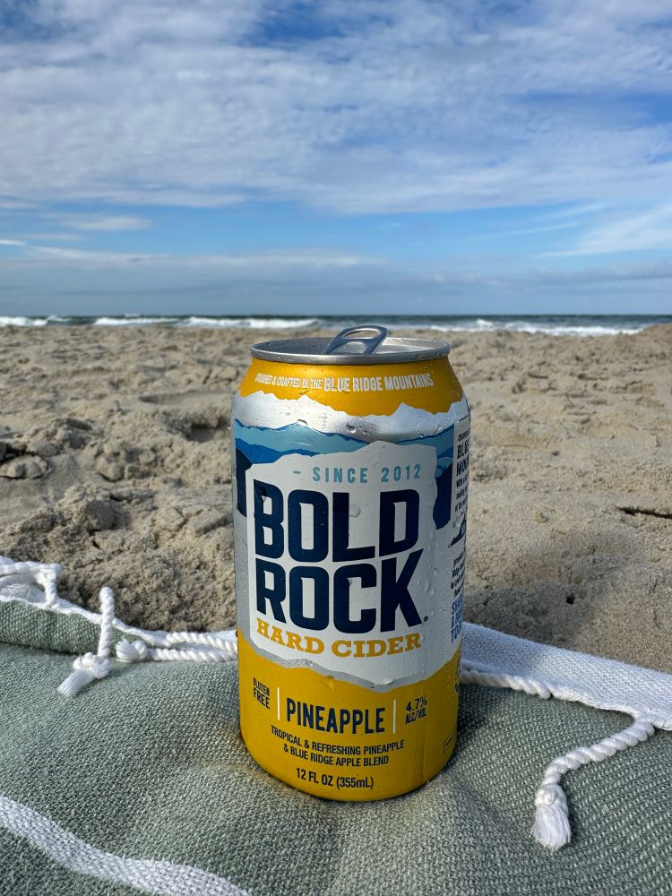 A bright yellow can of Bold Rock pineapple cider sitting on the edge of a pale green beach towel, sand and ocean waves behind it.