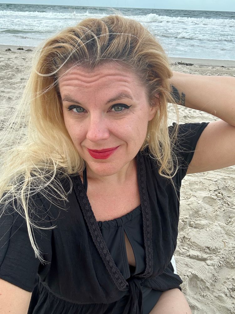 Cass looking heckin’ cute on the beach, a black cover-up draped over her black swimsuit, red lipstick, green eyes sparkling. Her hair is wind-tousled and held back from her face with one hand.