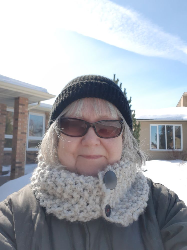 A woman wearing coat ,scarf, and toque, and sunglasses outside on a bright winter day.