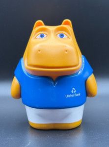 Photo of an old Henry Hippo money box, given to henry hippo childrens savings accounts from Ulster bank. It's an orange plastic hippo with blue eyes standing upright, wearing a blue top and white shorts. 