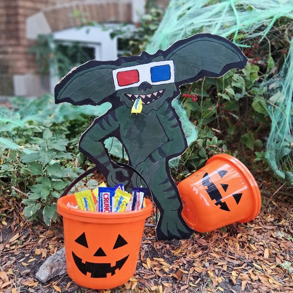 A plywood Gremlin with 3D glasses on holds a jack-o-lantern candy bucket full of candy wrappers. A wrapper hangs from his mouth.