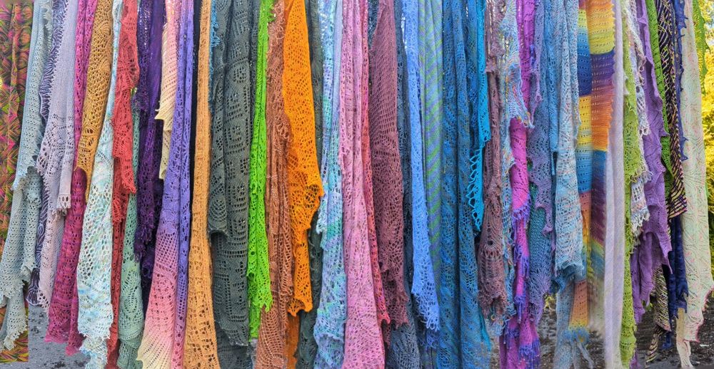 A panoramic photo showing many brightly coloured hand knitted lace shawls hanging down (from a washing line, but that's not in the photo)