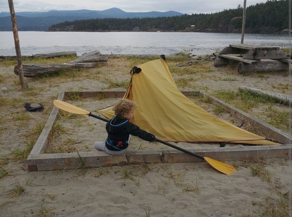 Small child sitting on the edge of a tent pad demonstrating kayak paddling technique in the sand. The tent is close to water. You can see near land across the bay and mainland in the distance. 