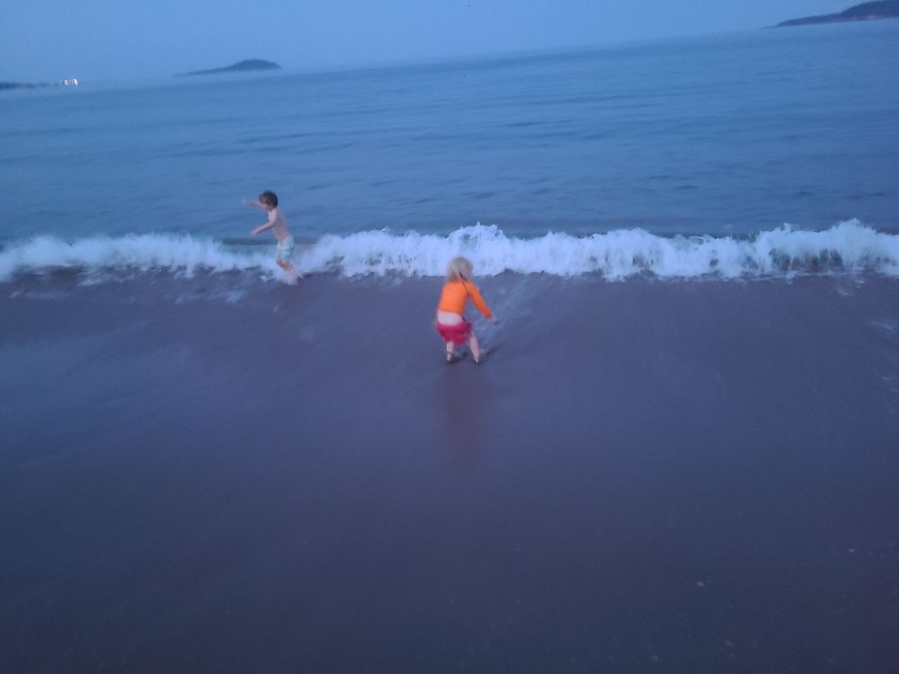 2 children playing in waves. One wearing orange and pink suit and the other wearing light blue. Islands are visible in the distance. 