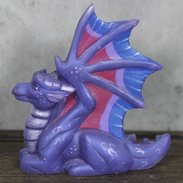 A figure of a purple baby dragon, loafing with his wings stretched out.  The wings are colored like the bisexual pride flag.