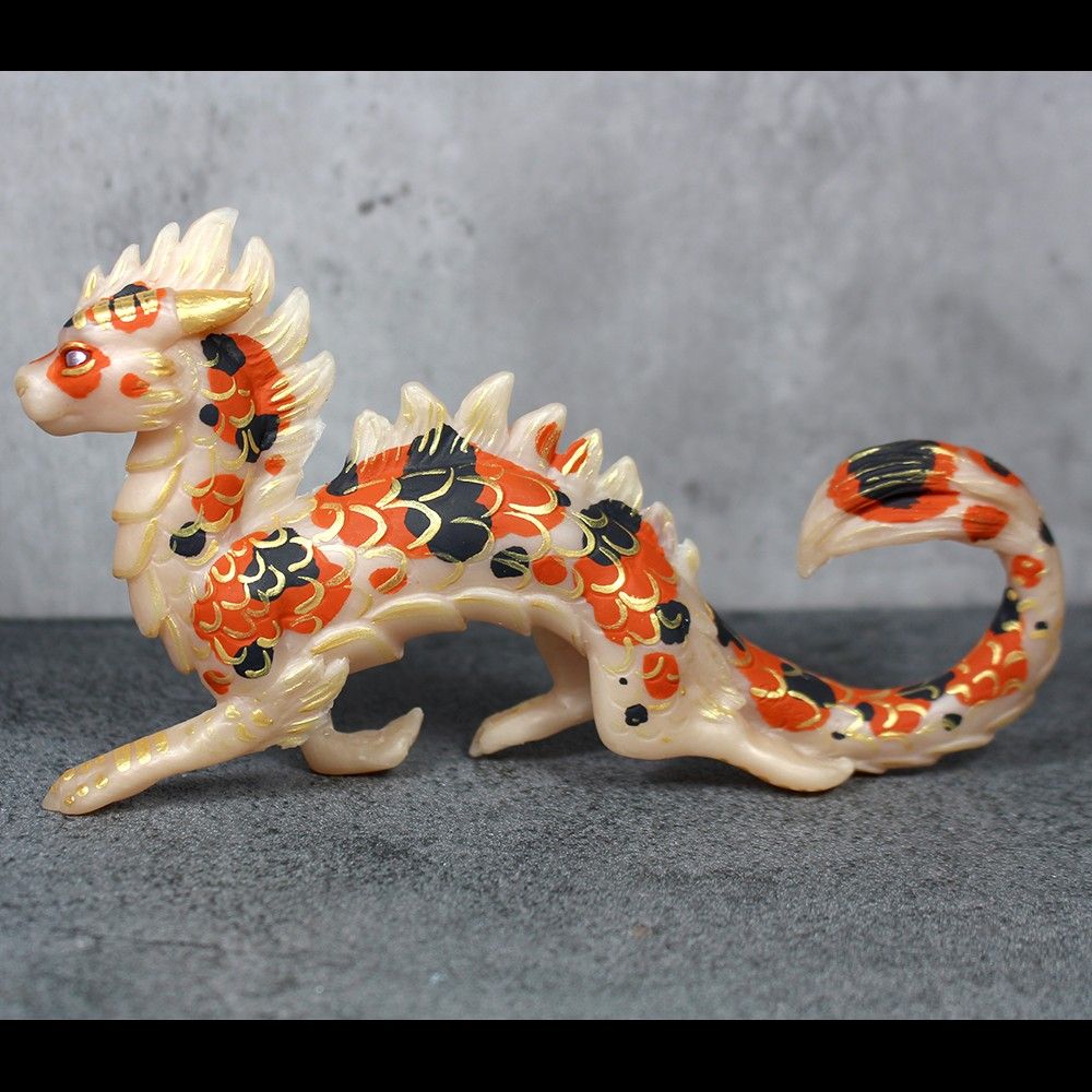 A sculpture of an eastern style dragon, painted like a koi fish.  He is prancing jauntily.  He is white with black and orange spots, accented with metallic gold scales.