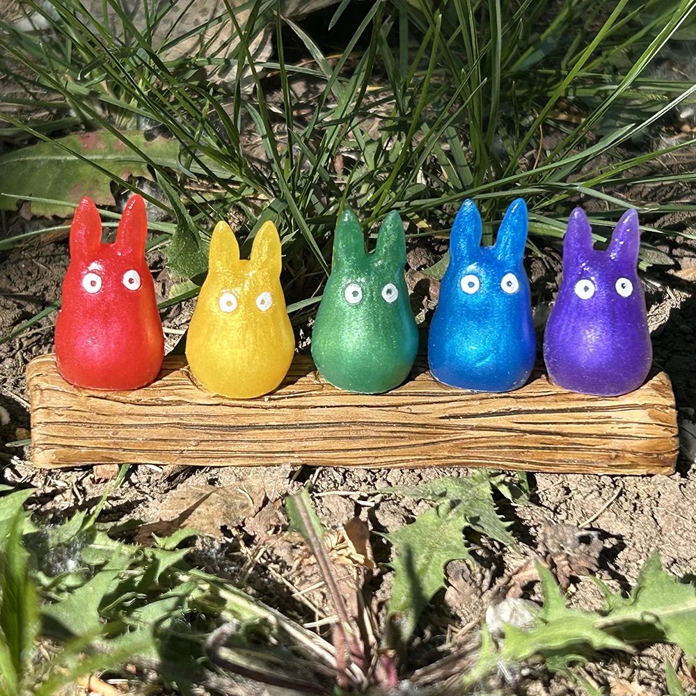 Five small totoro figures sitting on a log. They are rainbow colored on a grassy background.