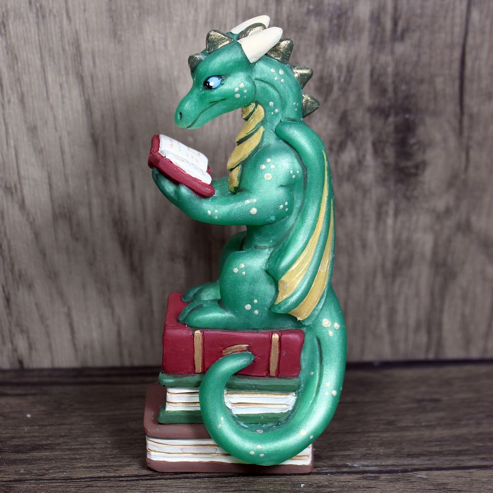 A figure of a green dragon sitting on a stack of books and reading a book of his own. 