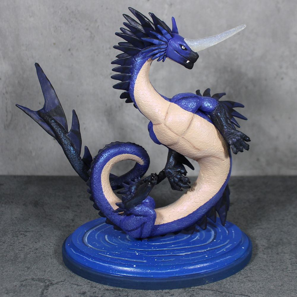 A sculpture of a long blue dragon with an array of back fins.  She has a long crystal horn on her head and hovers above a pool of water.  Front view.