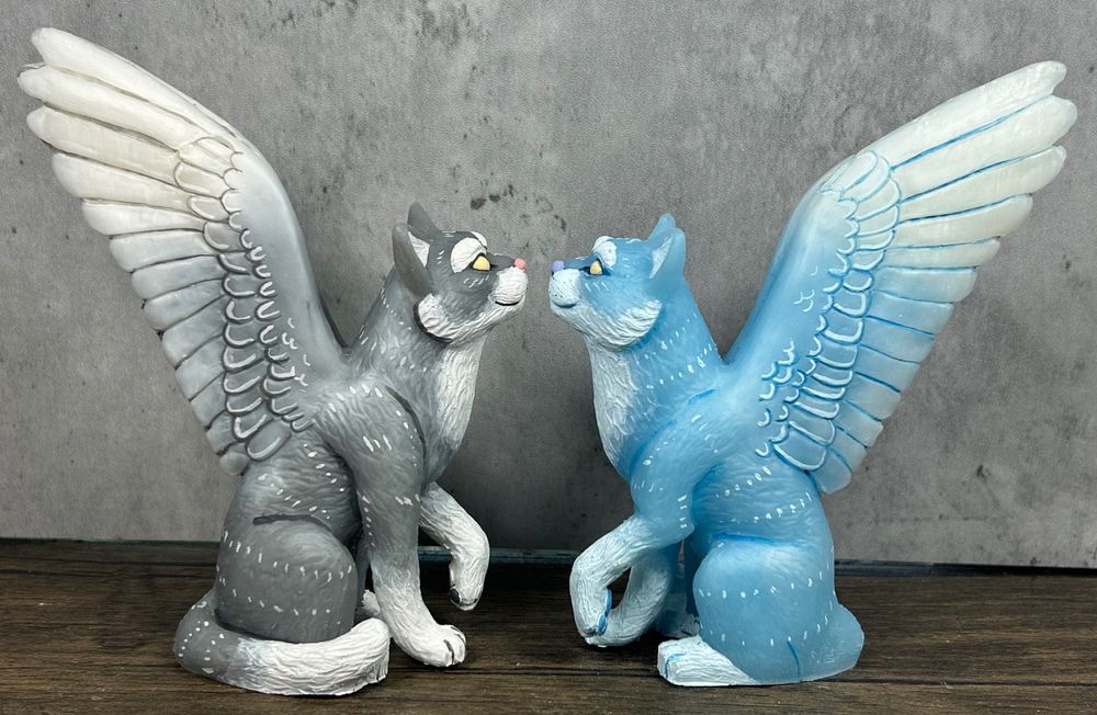 A photo of two winged cat figures sitting facing each other.  They have outstretched feathered wings.  One is grey and white and one is pale blue and white.