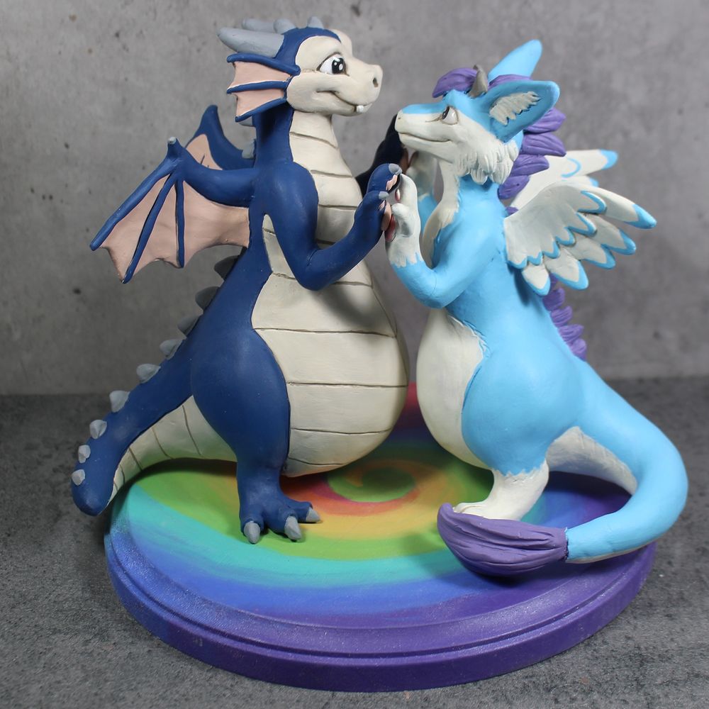 Two fat dragon figures stand facing each other with their paws together. They stand on a rainbow swirled base. 