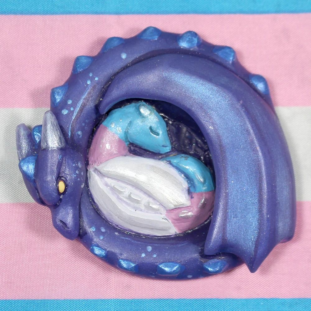 A sculpted magnet of a purple dragon curling protectively around a baby dragon colored like the trans flag. 