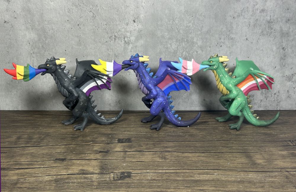A photo of three dragon figures. They are all rearing and breathing fire with their wings outspread. One is black with asexual pride wings and a rainbow flame. One is purple with bisexual pride wings and a non binary flame. One is green with lesbian pride wings and a trans flame. 