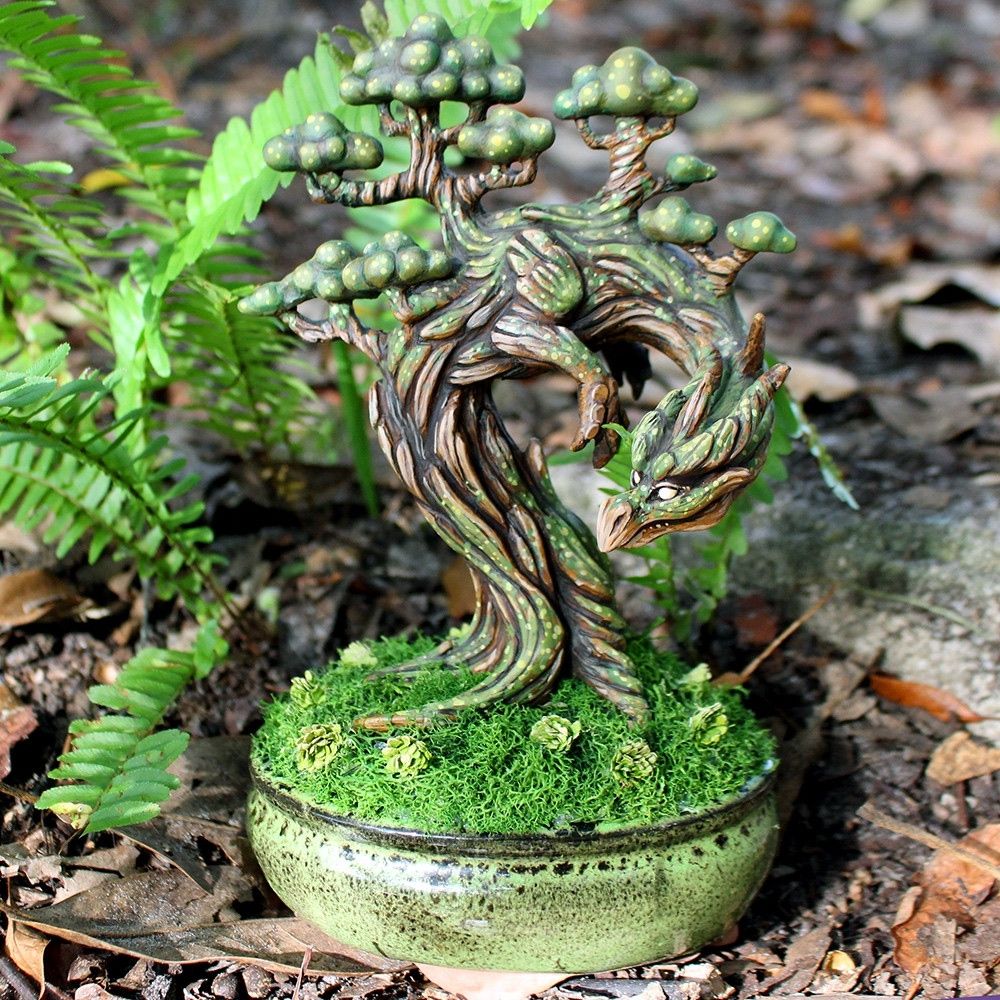 A tree dragon figure planted in a green bonsai pot. It is covered in green moss and its roots are surrounded by green reindeer moss. 
