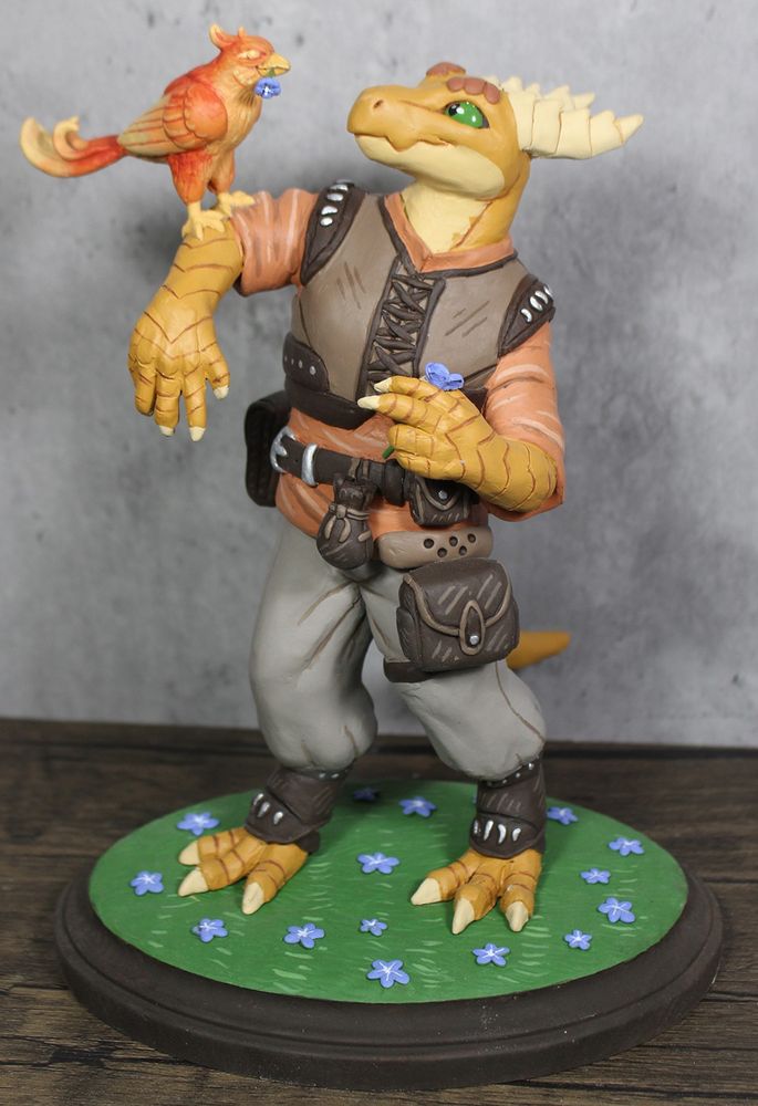 A dusty gold anthro dragon character. He is dressed in tan pants with an orange shirt and brown vest. He has various pouches attached to his belt.  He is holding his Phoenix friend on an outstretched arm. They are both smiling and looking at each other. Front view.