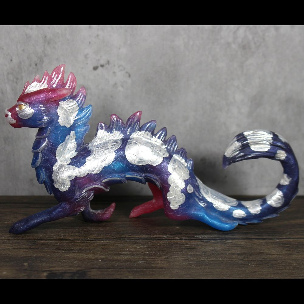 A sculpture of an eastern style dragon, painted like a koi fish.  He is prancing jauntily.  He is galaxy colored in purple/blue/pink metallic swirl with silver spots.  