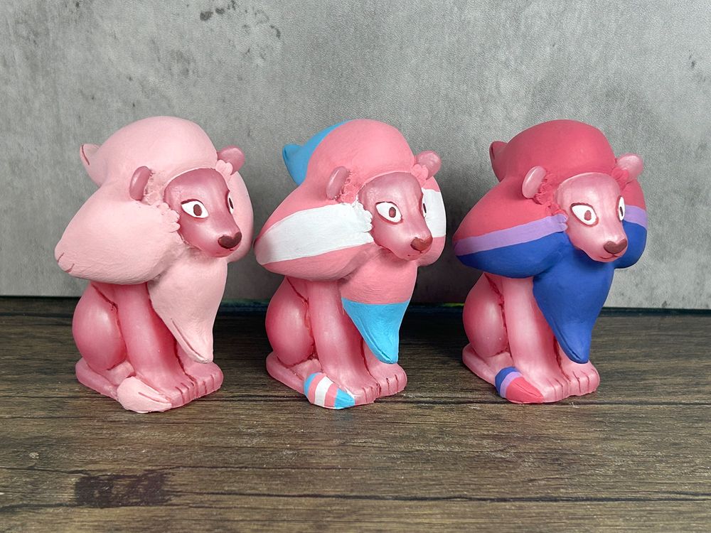 Three figures of Lion from Stephen Universe.  They are all sitting with their tails curled around them.  One is the classic pink coloration.  The second has a mane and tail tuft colored in the trans pride flag.  The last is colored in the bi pride flag.