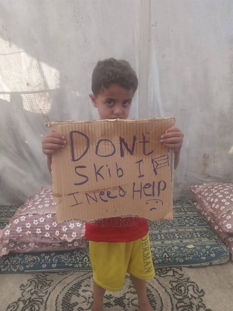 Young Muhammad stands in a tent holding a sign that reads “Don’t skip. I need help.”
