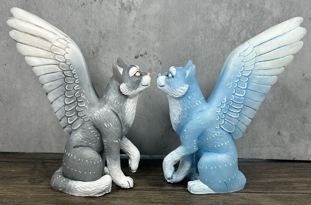 A photo of two winged cat figures sitting facing each other.  They have outstretched feathered wings.  One is grey and white and one is pale blue and white.
