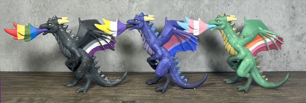 Three dragon figures - black, purple and green.  They are each rearing on their back legs, breathing fire.  The flames and wing membranes are pride flag colored.