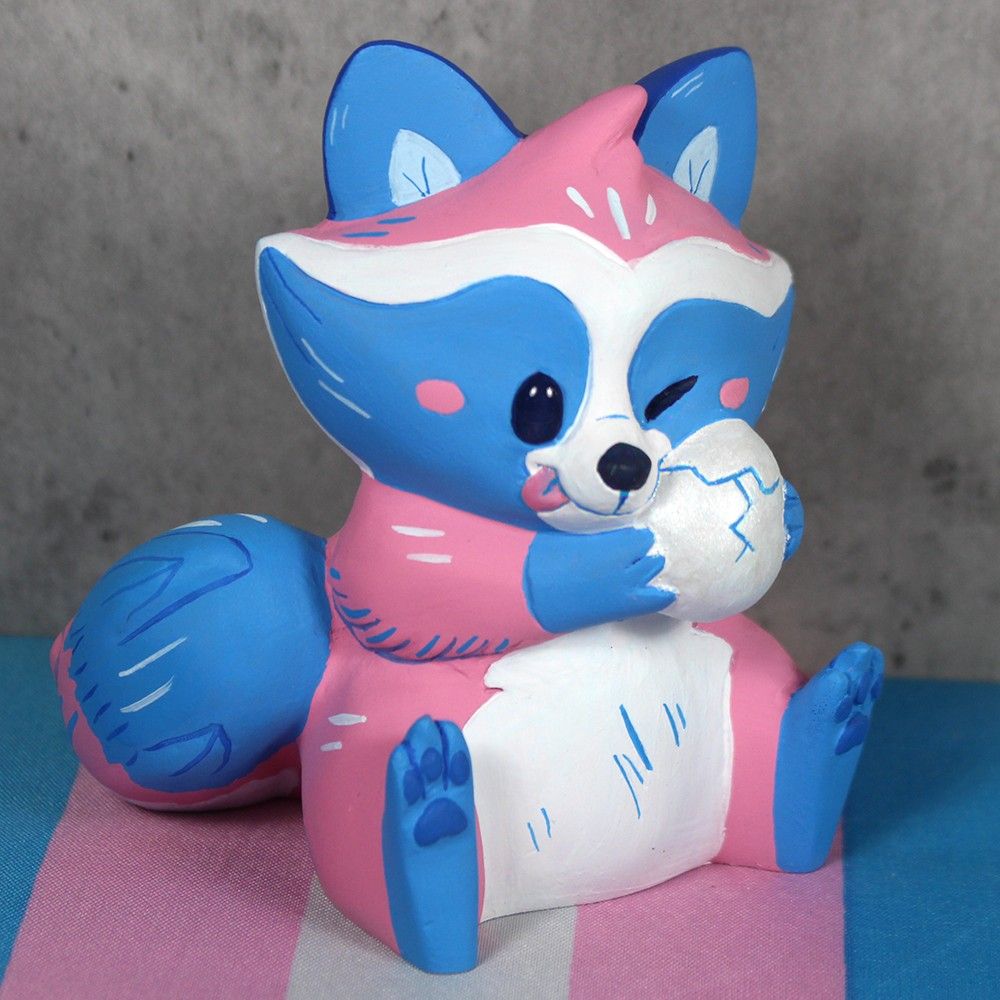 A cute sitting raccoon figure.  They are colored like the trans flag and are holding a cracked egg against their face.