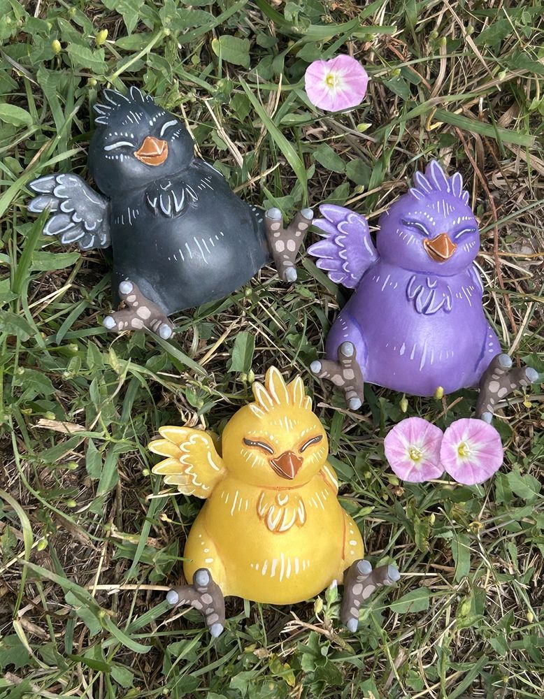 A photo of three fat chocobo figures lying in the grass.  One is yellow, one is lavender, and one is black.  There are a few pink flowers in the grass.