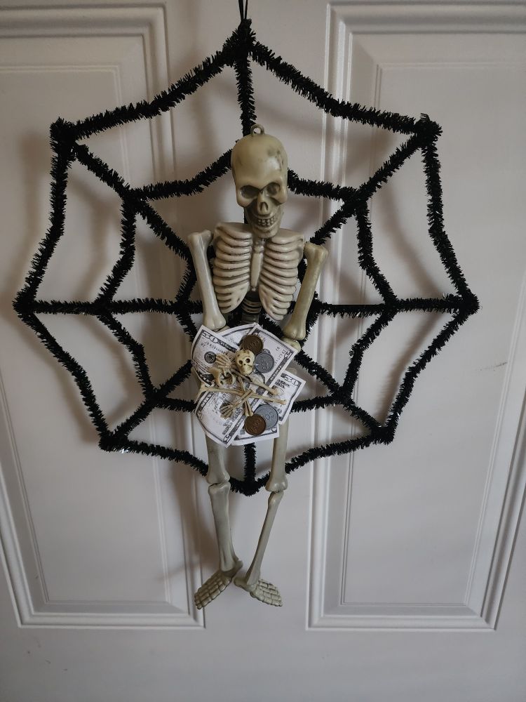 A Halloween wreath that is a spider web with a skeleton in the center ofit. The skeleton is holding a pile of money and bones. It is a reference to an "I Think You Should Leave" sketch that is beloved in our household.