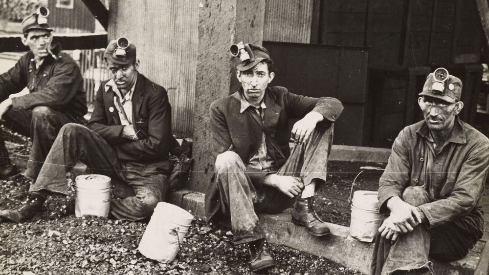 A black and white photo of 4 19th century coal miners sitting next to their buckets and wearing hats with headlamps attached. The point of the photo is to demonstrate that competitive eating world champion Joey Chestnut looks like he could fit right in 